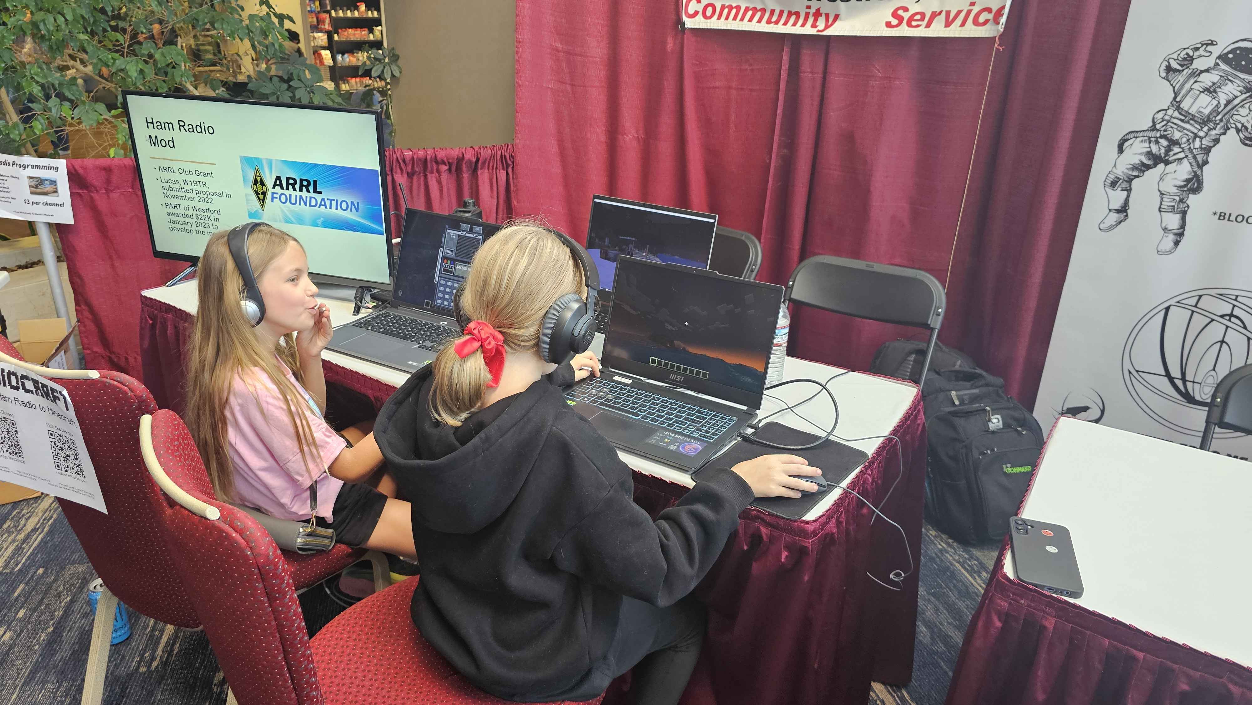 Two young attendees wearing headsets playing the Radiocraft Minecraft mod at the HamXposition booth with an ARRL Foundation presentation screen behind them